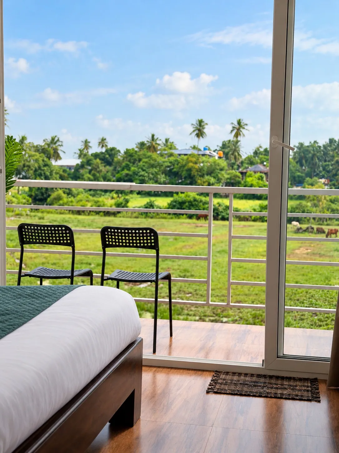 From the bed to the balcony: two chairs at the railing and green fields and palms beyond